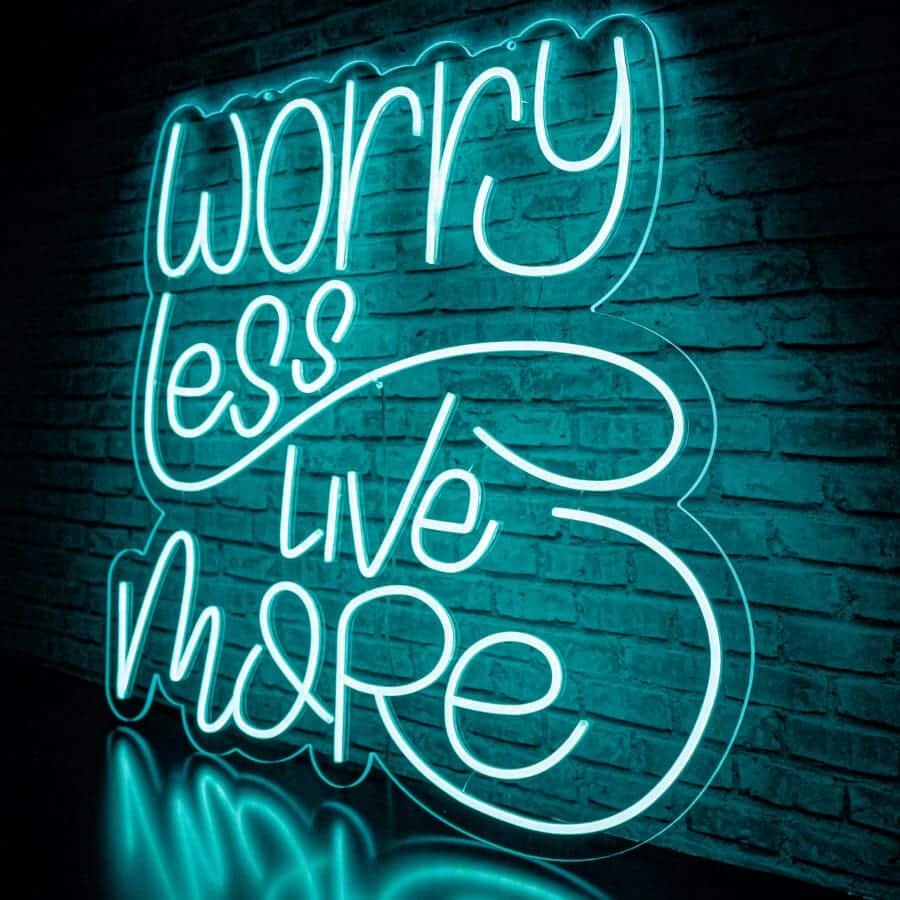 Néon "Worry Less Live More" sur un mur de briques avec la phrase "Préoccupe-toi moins, vis davantage" en lettres cursives et illuminé par des lumières bleu-vert.