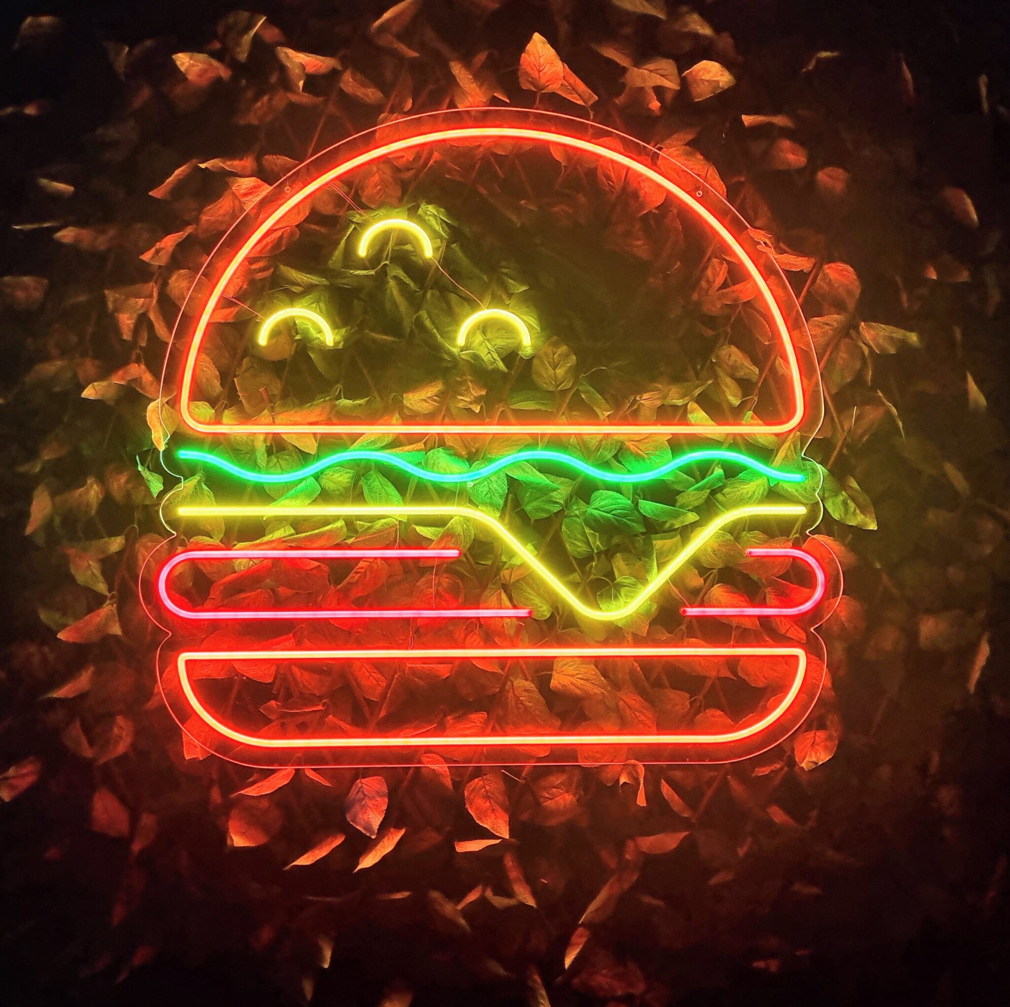 The Neon Burger sign glows brightly, featuring vibrant lettuce, cheese, and a patty against a background of dark leaves.