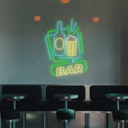 The Neon BAR sign features a bottle, glass, and the word "BAR," illuminating the dimly lit wall above empty tables and stools in this moody bar interior.