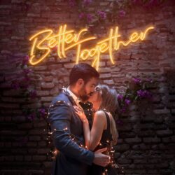 A couple in formal attire embraces and kisses, wrapped in string lights, standing before a brick wall illuminated by the Better Together Neon sign.