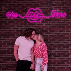 A couple kisses in front of a brick wall with the Kiss Me Here Neon sign glowing pink above a lip outline.