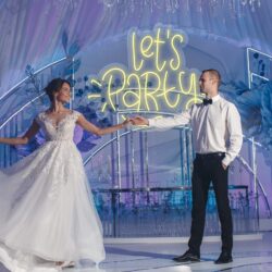 A bride and groom dance together under the Neon Let's Party sign at a beautifully decorated wedding venue.