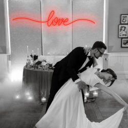 The bride and groom share a dance beneath the Neon Love sign, surrounded by candles and mist in a romantically decorated wedding venue.