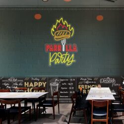The Neon Grill Party sign glows above empty restaurant tables and chairs, featuring a burger with flames, while a chalkboard-style menu is displayed below.
