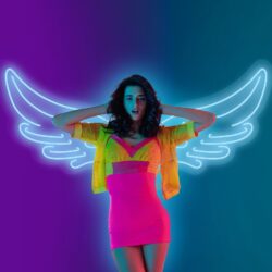 A woman poses in front of an Angel Wings Neon wall in a neon pink dress and yellow jacket, making it appear as if she has glowing wings behind her.