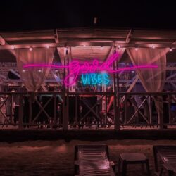 At night, the beachside bar features the Good Vibes Neon sign in pink and blue, glowing among lit string lights, sheer curtains, and empty lounge chairs in front.