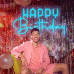 A man in a pink suit smiles in front of the Happy Birthday Neon sign, with balloons and shiny tinsel as festive decorations in the background.