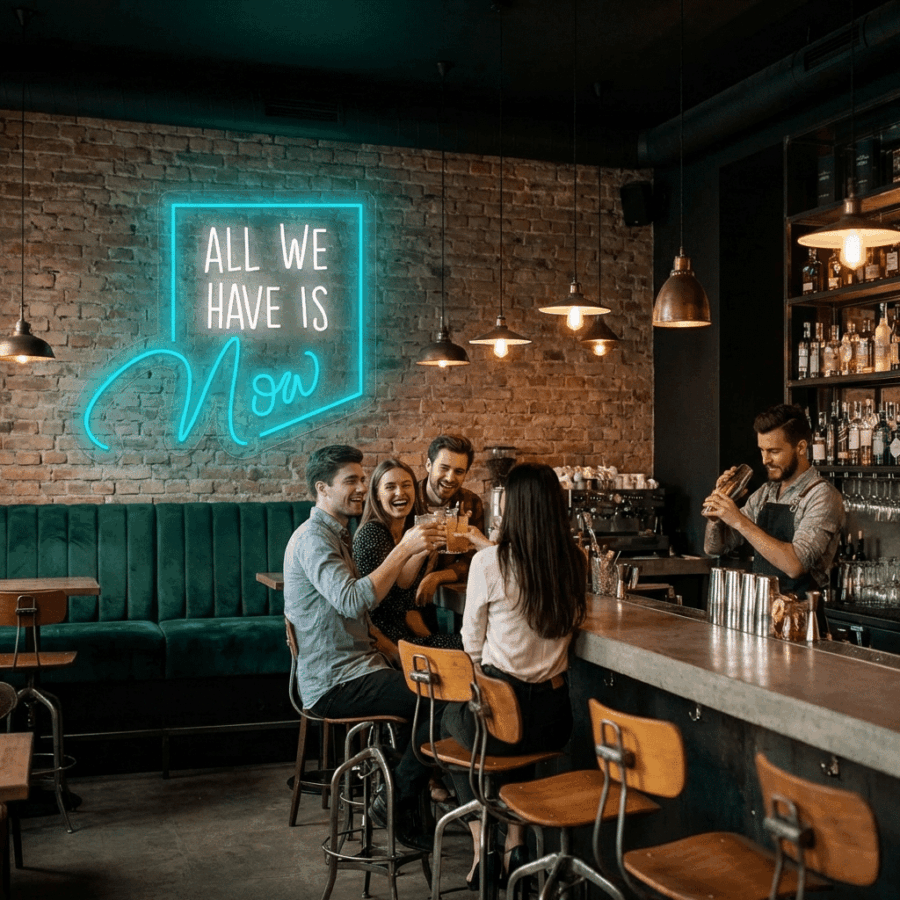 Four people sit at a bar counter with drinks, chatting and smiling, while a bartender prepares a cocktail. On the brick wall, the All we have is now neon casts a glow above them.