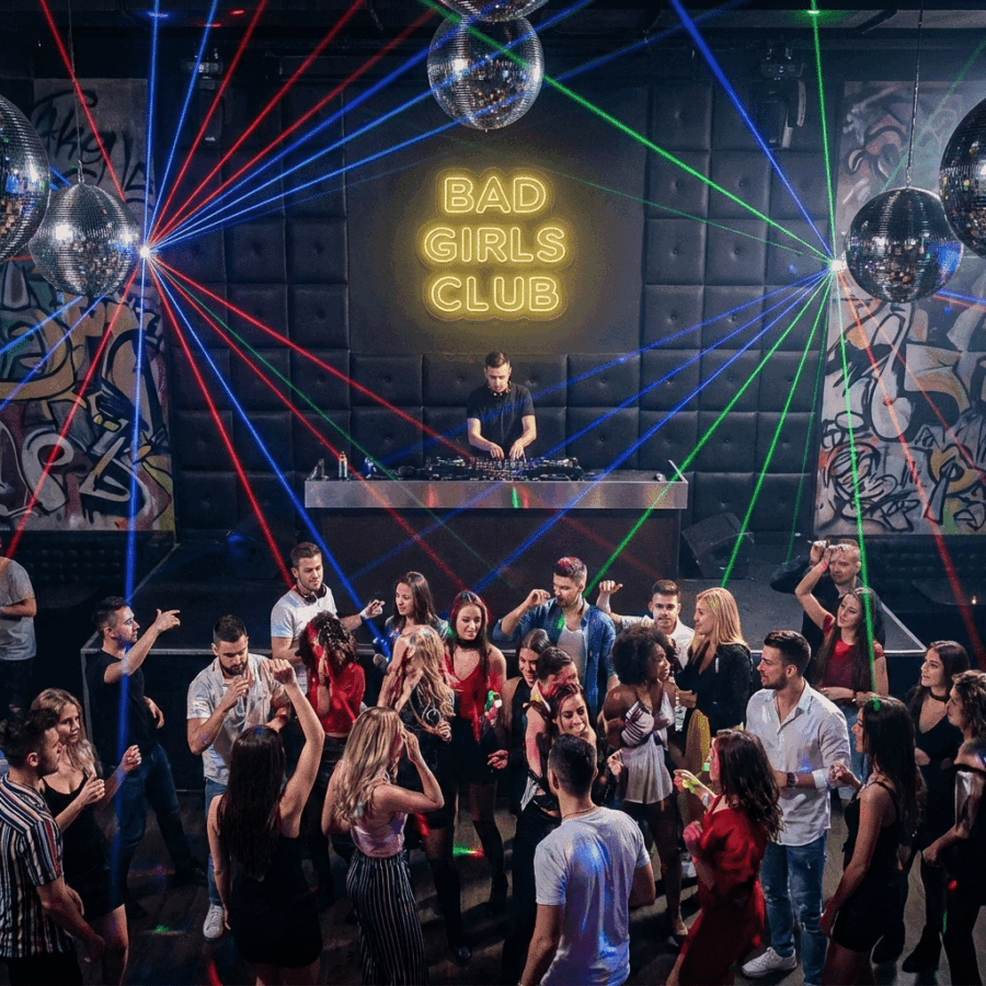 People dancing in a nightclub with colorful laser lights, as a DJ performs on stage beneath a glowing "Bad Girls Club Neon" sign.
