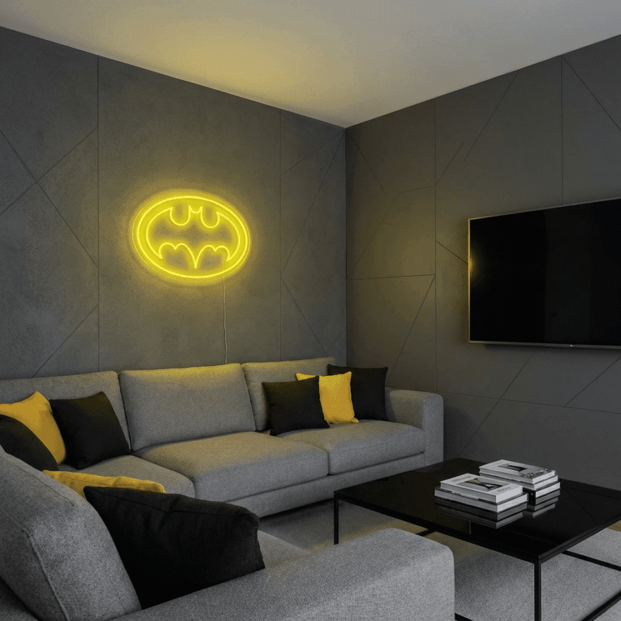 A modern living room with grey walls and furniture, black and yellow cushions, a black coffee table with books, and the Batman Neon light mounted on the wall.