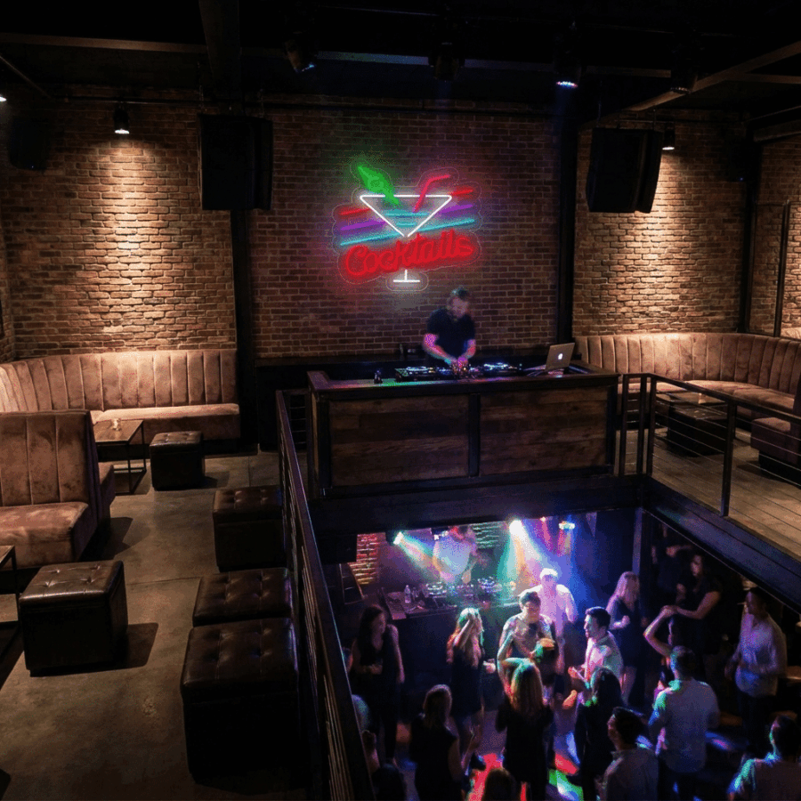 At Neon Cocktails, a DJ rocks an elevated booth amid brick walls and neon lights. Guests dance and snap selfies in vibrant light below, while the upper seating areas remain mostly empty.