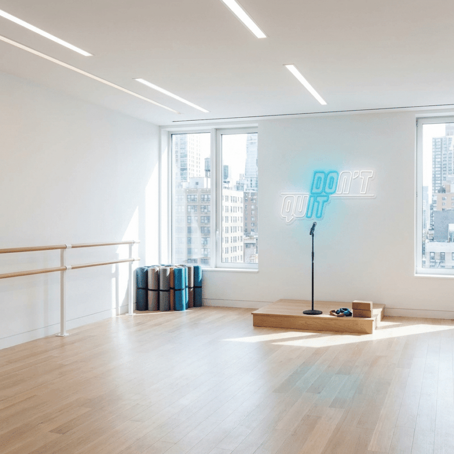 A bright dance studio with wooden floors, a barre on the left, stacked yoga mats, and a microphone on a small platform. The Neon Don't Quit sign glows on the wall while city buildings are visible outside.