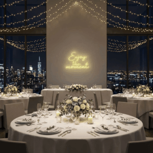 Round tables with white linens, floral centerpieces, candles, and string lights create an elegant dining room set for an event. The Neon Enjoy the Moment sign glows on the wall, enhancing the city skyline view at night.