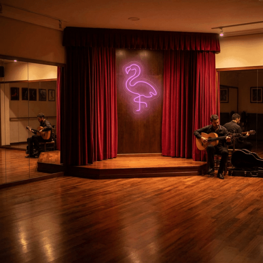 In a dimly lit room with wooden floors, two people play guitars beside a small stage adorned with red curtains and the bright Flamingo Neon sign.