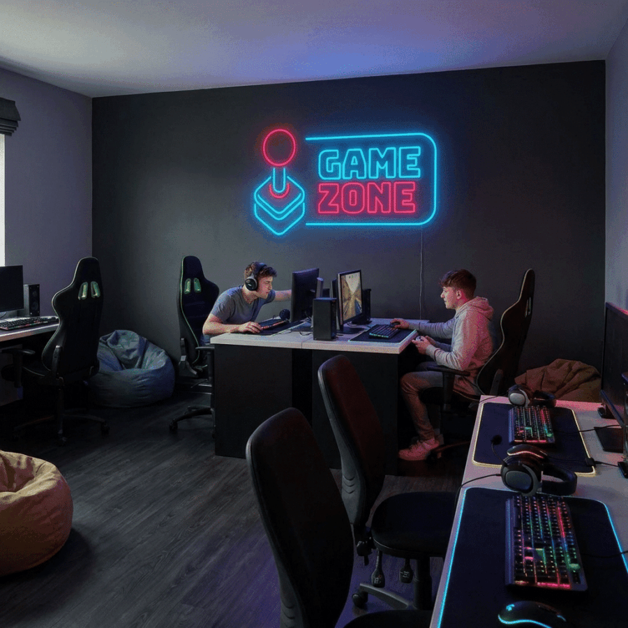 Two people sit at computers in a modern gaming room featuring the Neon Game Zone sign on the wall, surrounded by gaming chairs and keyboards.
