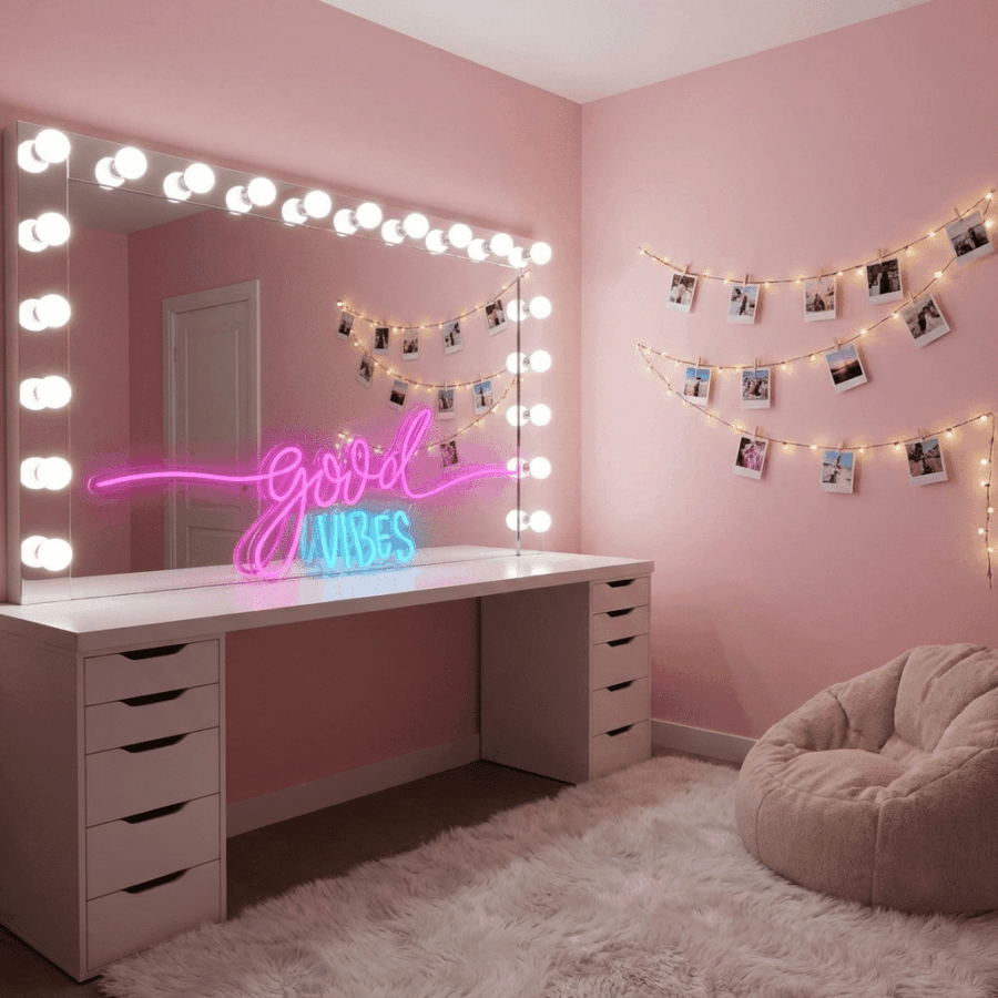 A pink room featuring a lighted vanity mirror, Good Vibes Neon sign, photo string lights on the wall, a white shag rug, and a beige bean bag chair.