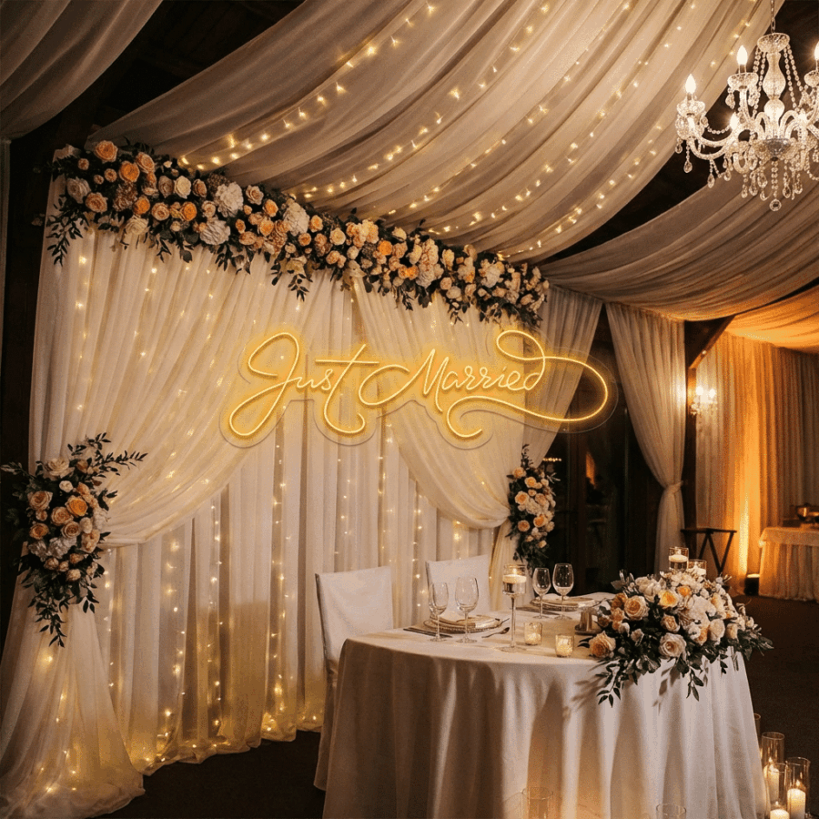 A wedding reception table decorated with flowers and glassware sits in front of draped white curtains with fairy lights and a Just Married Neon sign.