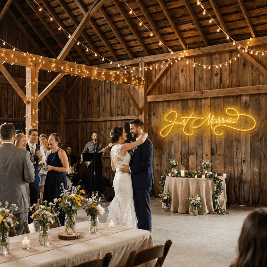 A bride and groom share their first dance in a rustic barn adorned with string lights and the Just Married Neon, while guests watch and a live band plays in the background.