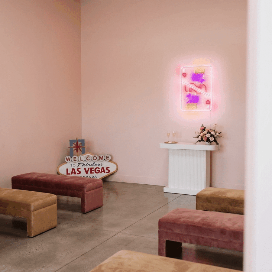 Minimalist room with pink walls, velvet benches, Neon K hearts sign, "Welcome to Las Vegas" sign, a small table with flowers, and two champagne glasses.