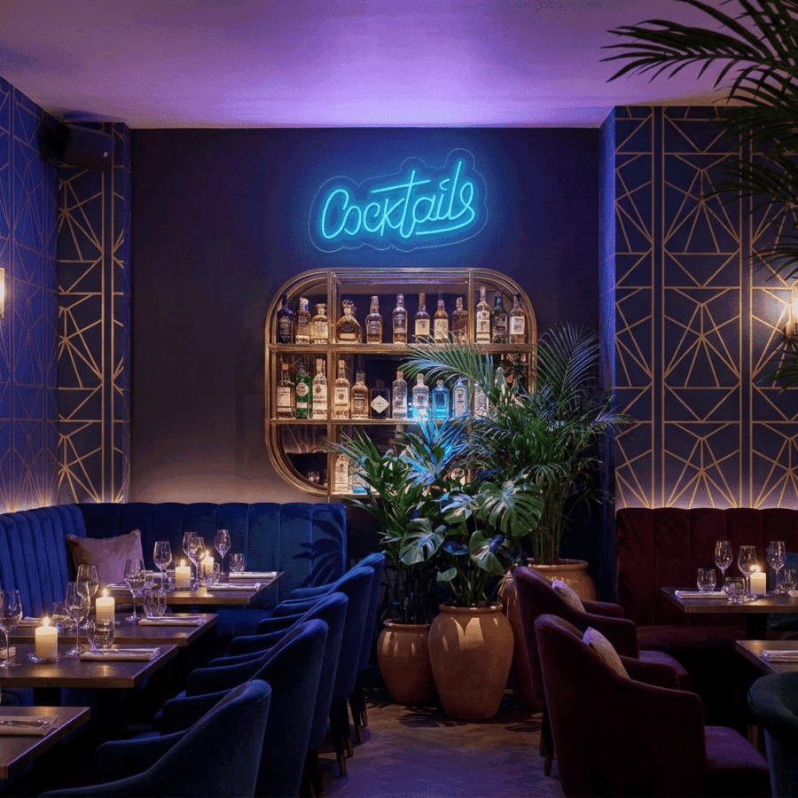 A stylish bar with blue and gold decor, candlelit tables, potted plants, and a neon "COCKTAILS" sign above a shelf of liquor bottles.