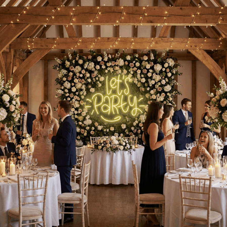 Wedding reception with guests mingling at round tables, large floral centerpieces, and the Neon Let's Party sign on a flower wall backdrop. Warm lighting and wooden beams add charm to the scene.