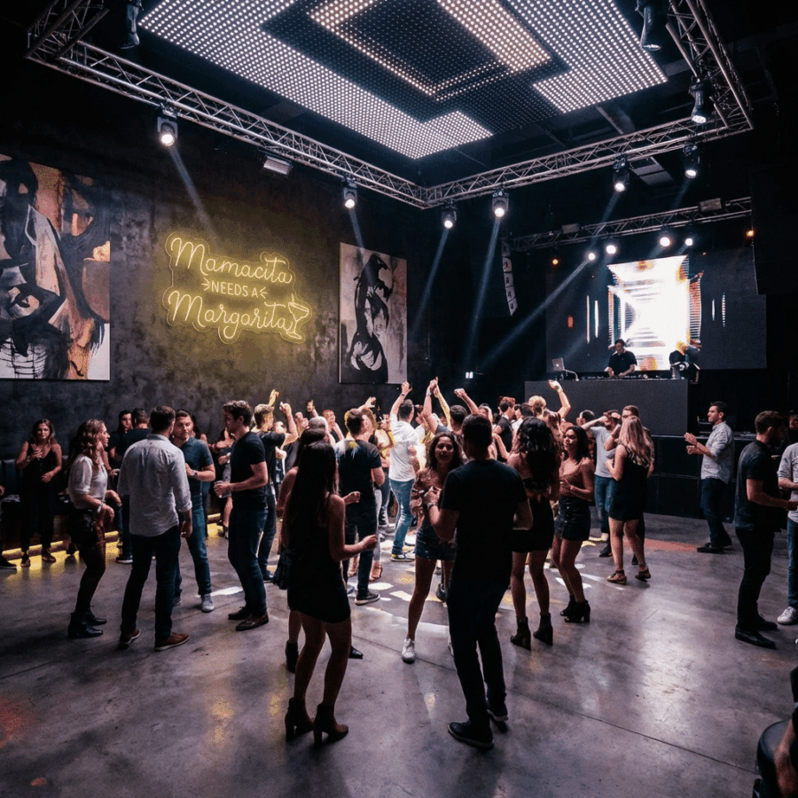 A crowd dances in a nightclub with a DJ on stage. The neon "Mamasita Needs a Margarita" sign glows, surrounded by modern lighting and art that energize the lively space.