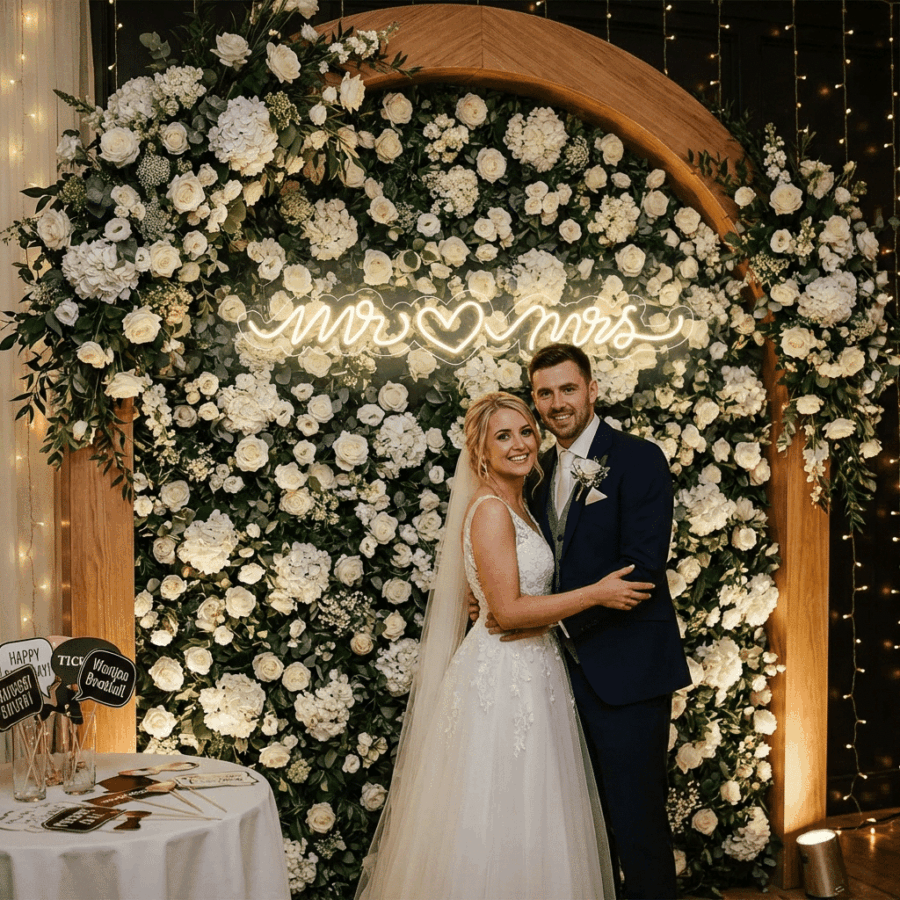 A bride and groom smile in front of a large floral arch featuring the Mr & Mrs Neon sign at their wedding reception.