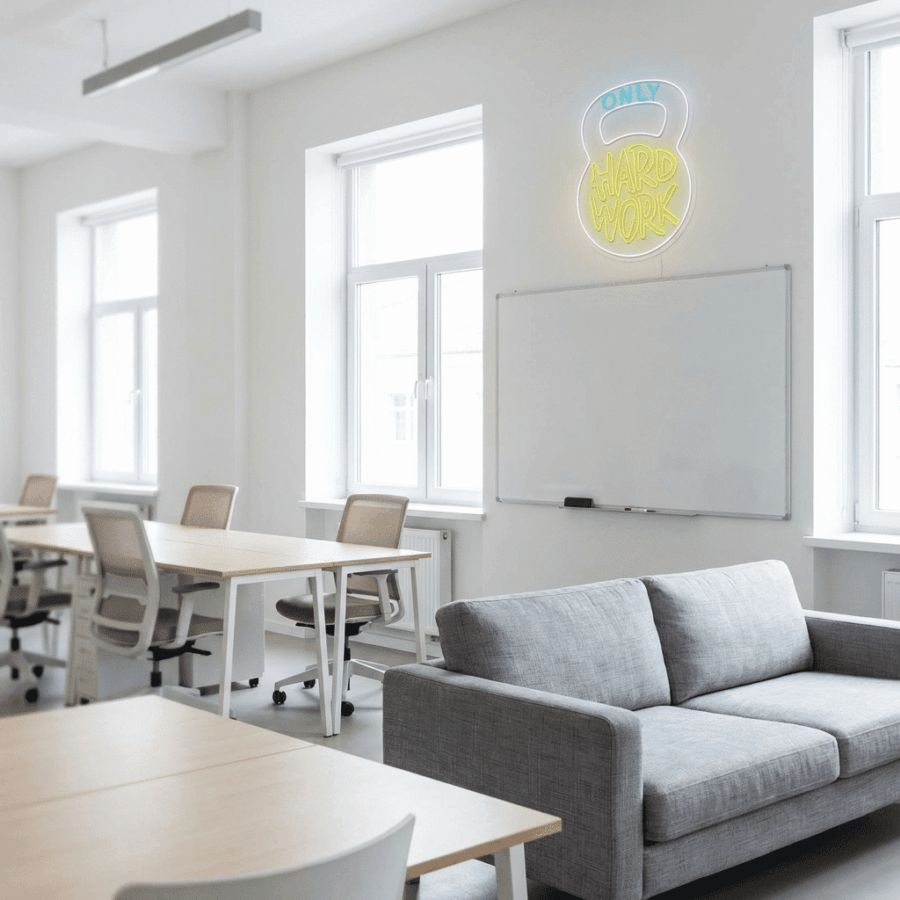 A modern office with white walls, desks, chairs, a gray sofa, and a whiteboard. Above the whiteboard is the Neon Only Hard Work sign. Natural light streams through large windows.