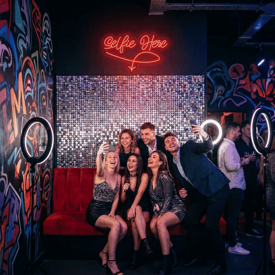 Six people pose for selfies on a red couch in a graffiti-decorated room lit by two ring lights and the Selfie Neon Here sign, with additional people visible in the background.