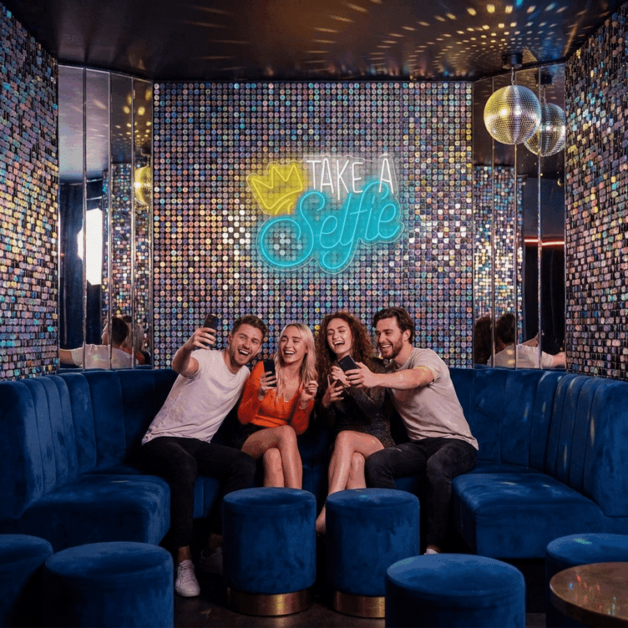 Four people sit on a blue velvet couch in a nightclub, taking a selfie under a disco ball. Behind them, the vibrant wall features the glowing Neon Take a Selfie sign, enhancing the lively vibe.