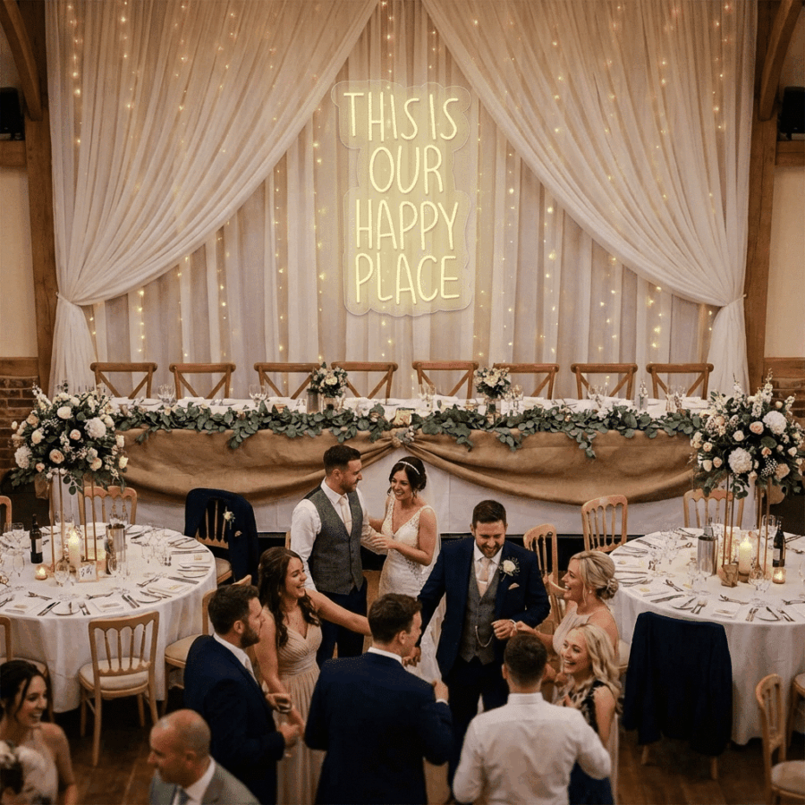 Guests dance at a wedding reception in front of a decorated head table, beautifully lit by the Neon This is our Happy Place sign.