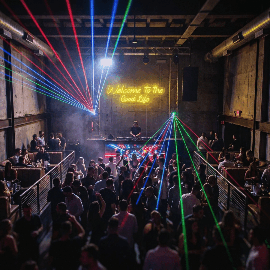 A lively nightclub scene features people dancing, vibrant laser lights, and a DJ on stage beneath the Neón Welcome to the Good Life sign as guests take selfies to capture the energetic atmosphere.