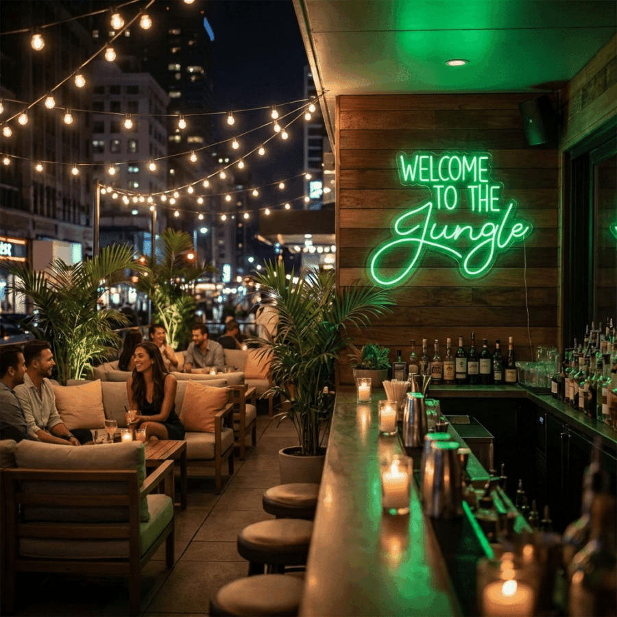 At night, people relax on couches under string lights and a glowing Welcome To The Jungle Neon Sign, surrounded by potted plants and a stocked bar counter in the outdoor lounge.