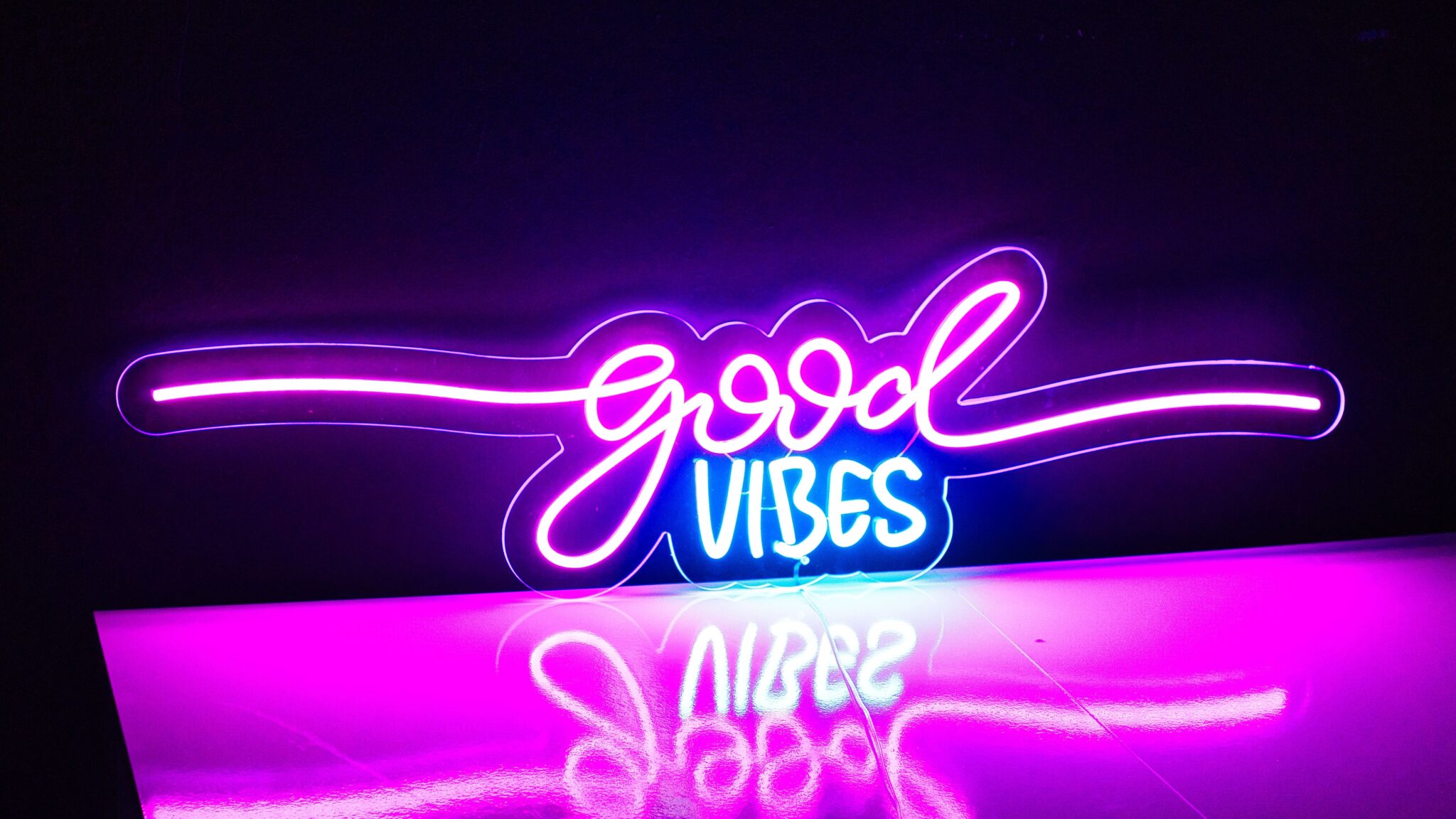 The Good Vibes Neon sign glows in pink, purple, and blue against a dark background, with its vibrant light reflecting on the shiny surface below.