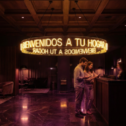 Three people stand at a reception desk in a dimly lit lobby with a large circular neon sign overhead reading "Bienvenidos a tu hogar.
